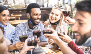Friends toasting red wine at outdoor restaurant bar with open face mask – New normal lifestyle concept with happy people having fun together on warm filter – Focus on afroamerican guy